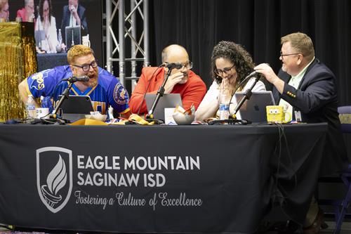 Three people whisper while a fourth yells at them behind a table